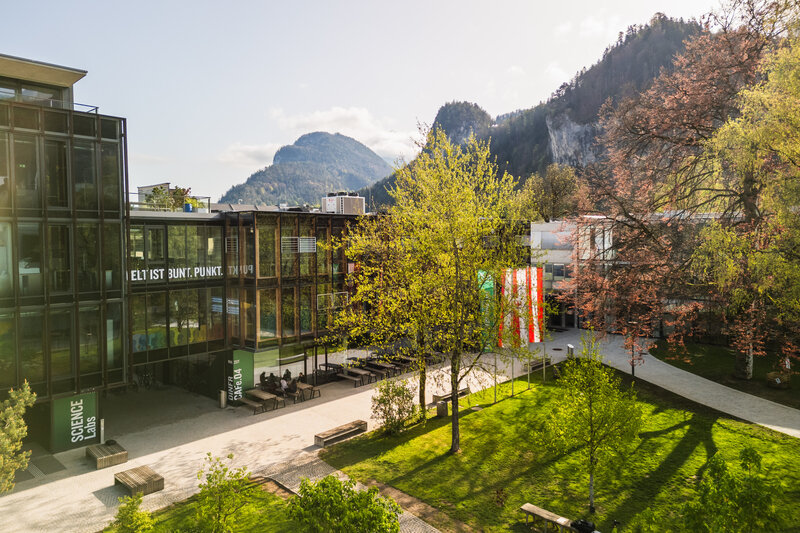 FH Kufstein Tirol – moderner Hochschulcampus mit Alpenblick Frühlingshafte Außenaufnahme des Campus der FH Kufstein Tirol mit Glasfassade, Fahnen und Bergkulisse im Hintergrund