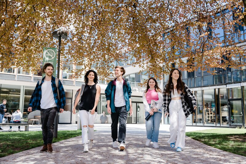 Gemeinsam studieren, gemeinsam wachsen – Campusleben an der FH Kufstein Tirol Fünf Studierende gehen gemeinsam über den Campus bei Frühlingswetter
