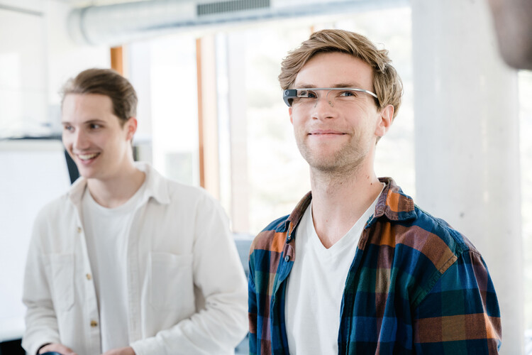 Zwei Männer in einem hellen Raum. Der vordere trägt eine Eye-Tracking Brille und ein kariertes Hemd, der hintere ein weißes Hemd und lächelt. | © FH Kufstein Tirol