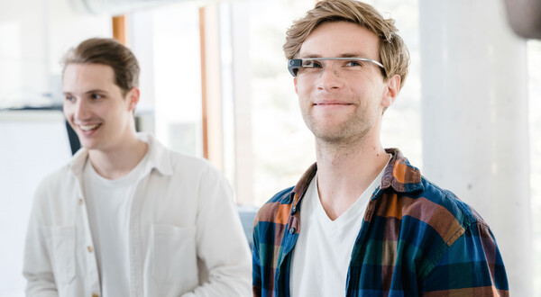 Zwei Männer in einem hellen Raum. Der vordere trägt eine Eye-Tracking Brille und ein kariertes Hemd, der hintere ein weißes Hemd und lächelt. | © FH Kufstein Tirol