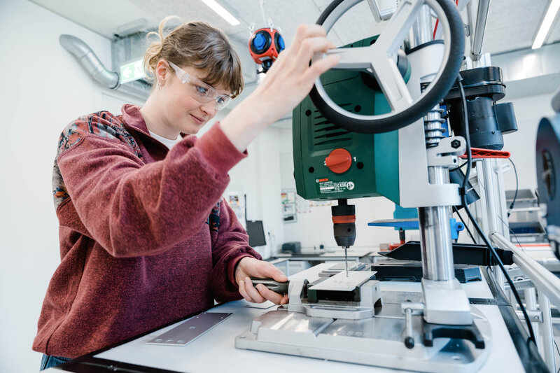 Eine Frau trägt eine Schutzbrille und arbeitet konzentriert mit einer stationären Bohrmaschine. | © FH Kufstein Tirol