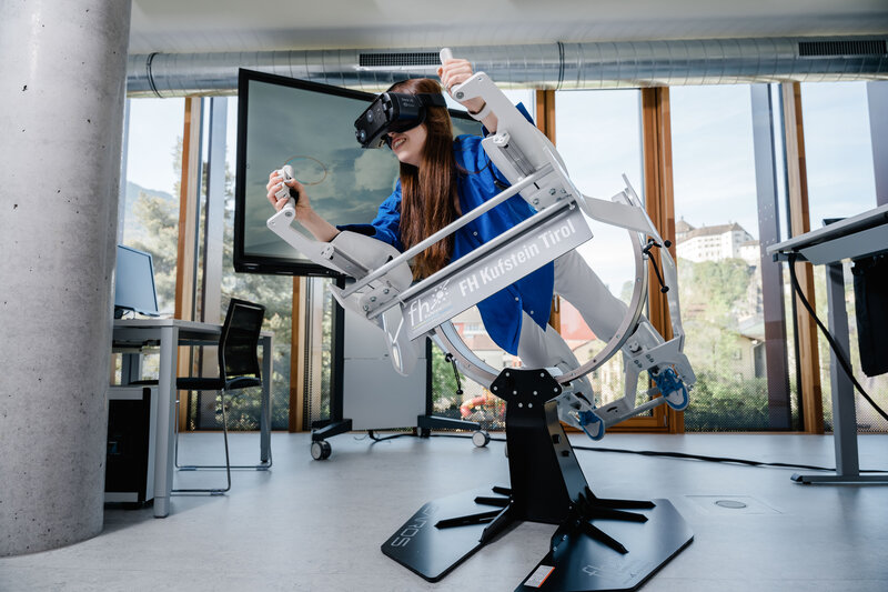 Eine Frau bedient eine VR Simulationsmaschine und liegt in einer Position, als ob sie fliegen würde. | © FH Kufstein Tirol