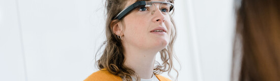 Eine Frau mit brünetten, lockigen Haaren trägt eine Eye-Tracking Brille und schaut leicht nach oben. | © FH Kufstein Tirol