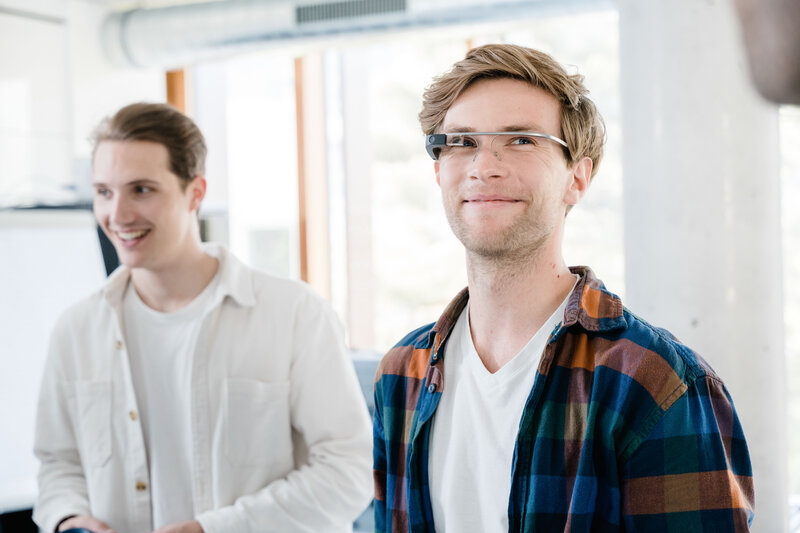 Zwei Männer in einem hellen Raum. Der vordere trägt eine Eye-Tracking Brille und ein kariertes Hemd, der hintere ein weißes Hemd und lächelt. | © FH Kufstein Tirol