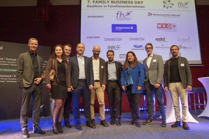 Neun Personen stehen auf einer Bühne vor einem Hintergrund, der mit Logos von Sponsoren und Partnern des ‘7. Family Business Day’ geschmückt ist. | © FH Kufstein Tirol