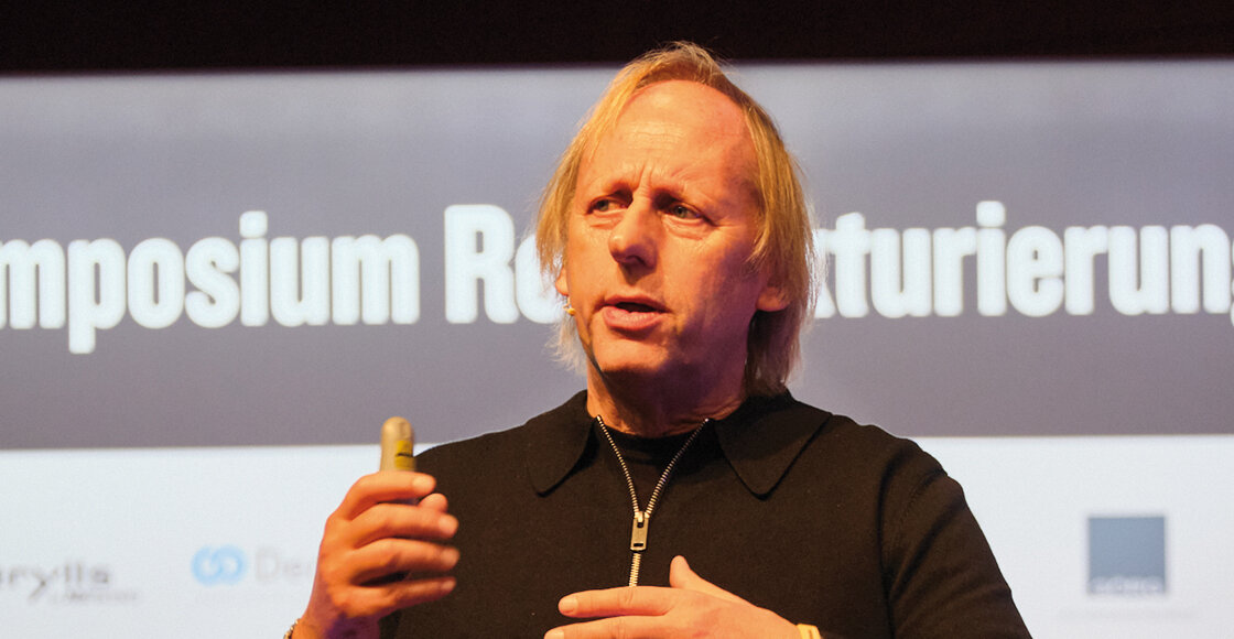 Ein Mann spricht auf einer Bühne und hält ein Präsentationsgerät in der Hand, im Hintergrund sind der Schriftzug „Symposium Restrukturierung“ und einige Sponsorenlogos zu sehen. | © FH Kufstein Tirol