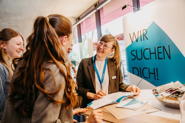 Drei Frauen im Gespräch an einem Messestand mit der Aufschrift "Wir suchen Dich!" im Hintergrund. | © FH Kufstein Tirol