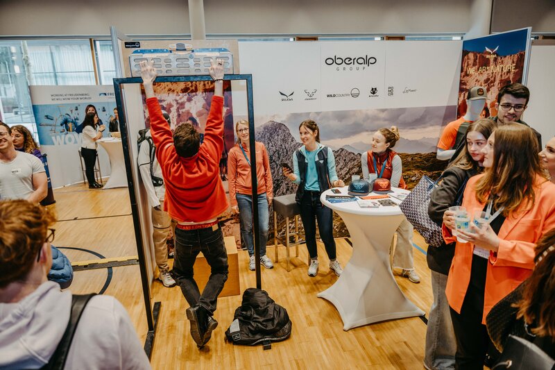 Ein Mann macht Klimmzüge an einem Klettergriffbrett vor dem Oberalp Messestand, während andere Besucher zuschauen und interagieren. | © FH Kufstein Tirol