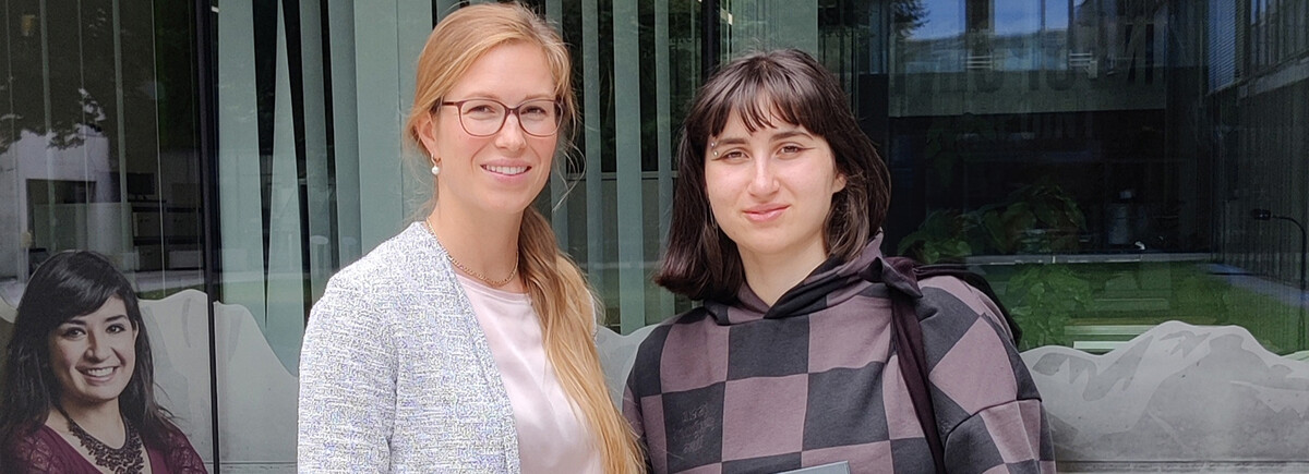 Zwei Frauen stehen nebeneinander und lächeln in die Kamera.  Im Hintergrund ist eine Glasfront mit einer spiegelnden Oberfläche zu sehen. | © FH Kufstein Tirol