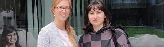 Zwei Frauen stehen nebeneinander und lächeln in die Kamera.  Im Hintergrund ist eine Glasfront mit einer spiegelnden Oberfläche zu sehen. | © FH Kufstein Tirol