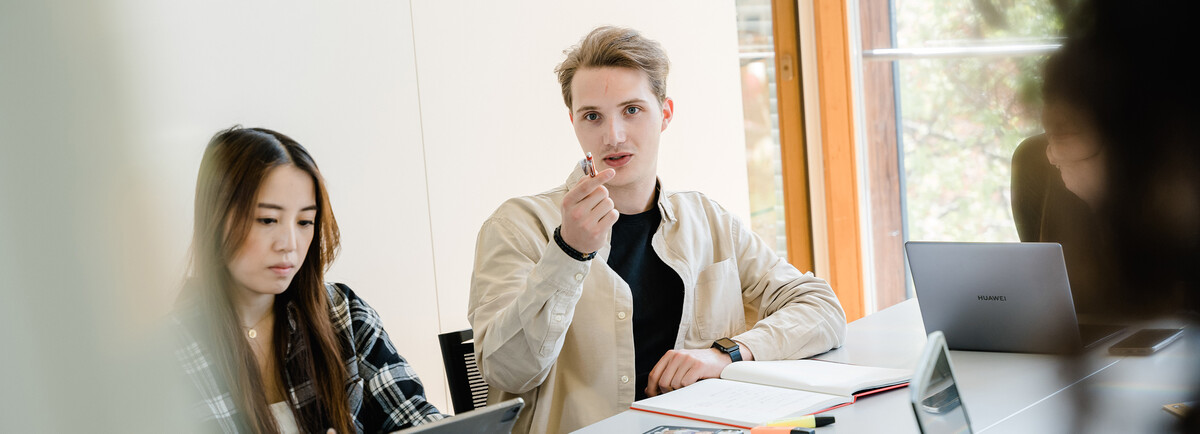 Drei Personen sitzen an einem Tisch, und ein Mann dieser Gruppe zeigt mit seinem Stift in die Richtung der Kamera, wo sich ein Whiteboard befindet. | © FH Kufstein Tirol