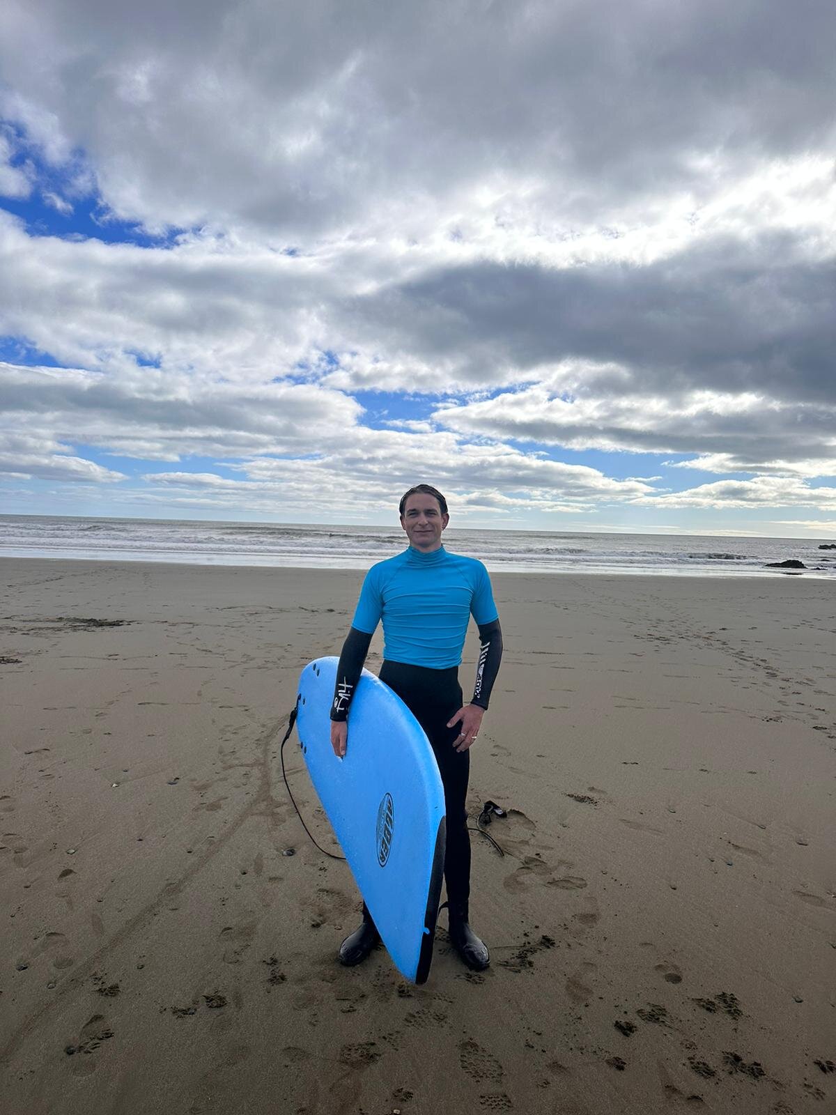 Ein junger Mann in einem Neoprenanzug und blauem Shirt mit einem Surfboard am Strand. | © Andreas Schmieger