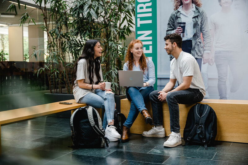 Drei Personen sitzen zusammen in der Aula und besprechen etwas freudig miteinander. | © FH Kufstein Tirol