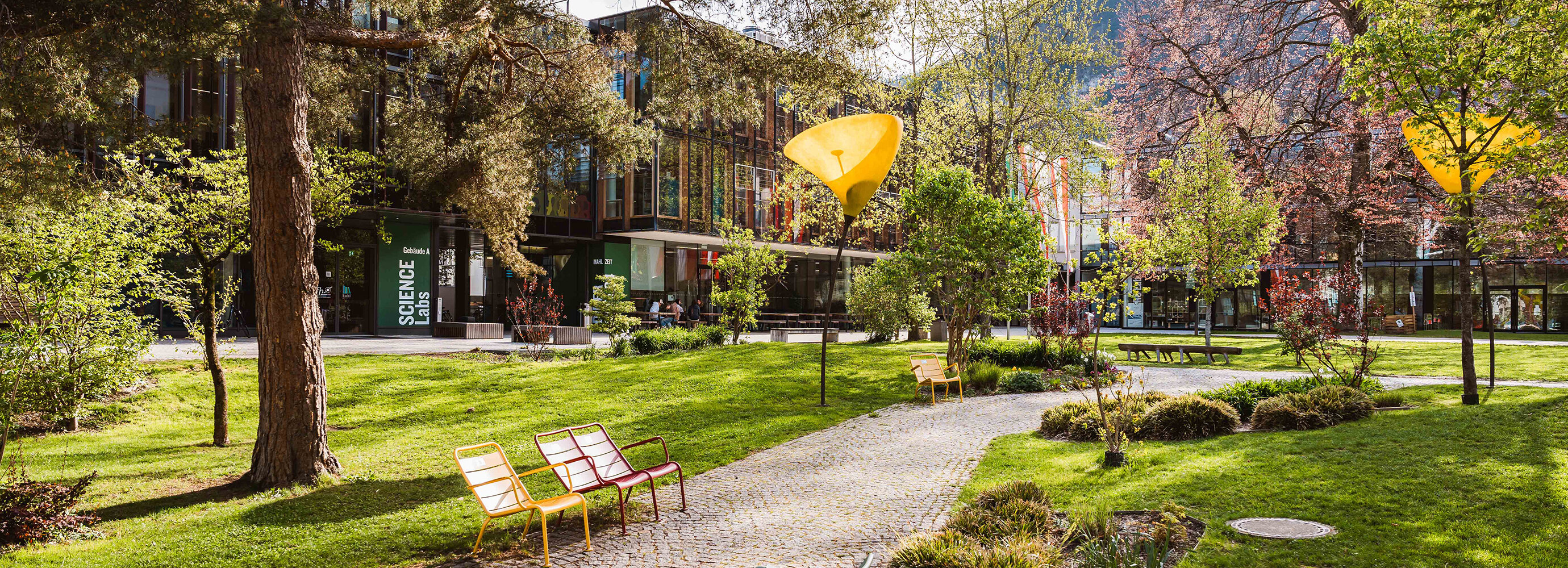 Grüner Park mit Wegen, bunten Sitzbänken und auffälligen gelben Lampen vor der FH Kufstein Tirol | © FH Kufstein Tirol