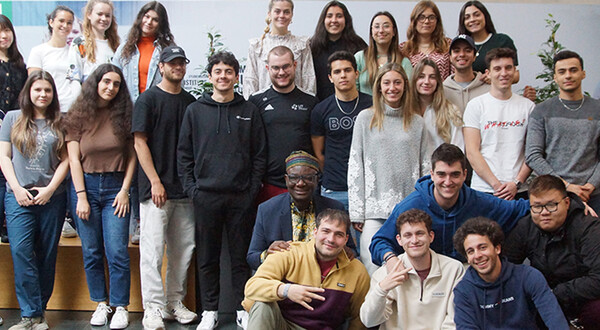 Eine Gruppe von Studierenden der FH Kufstein Tirol steht und sitzt lächelnd zusammen mit Gastlektor Bella Bello Bitugu in der Aula der Fachhochschule Kufstein Tirol. | © FH Kufstein Tirol