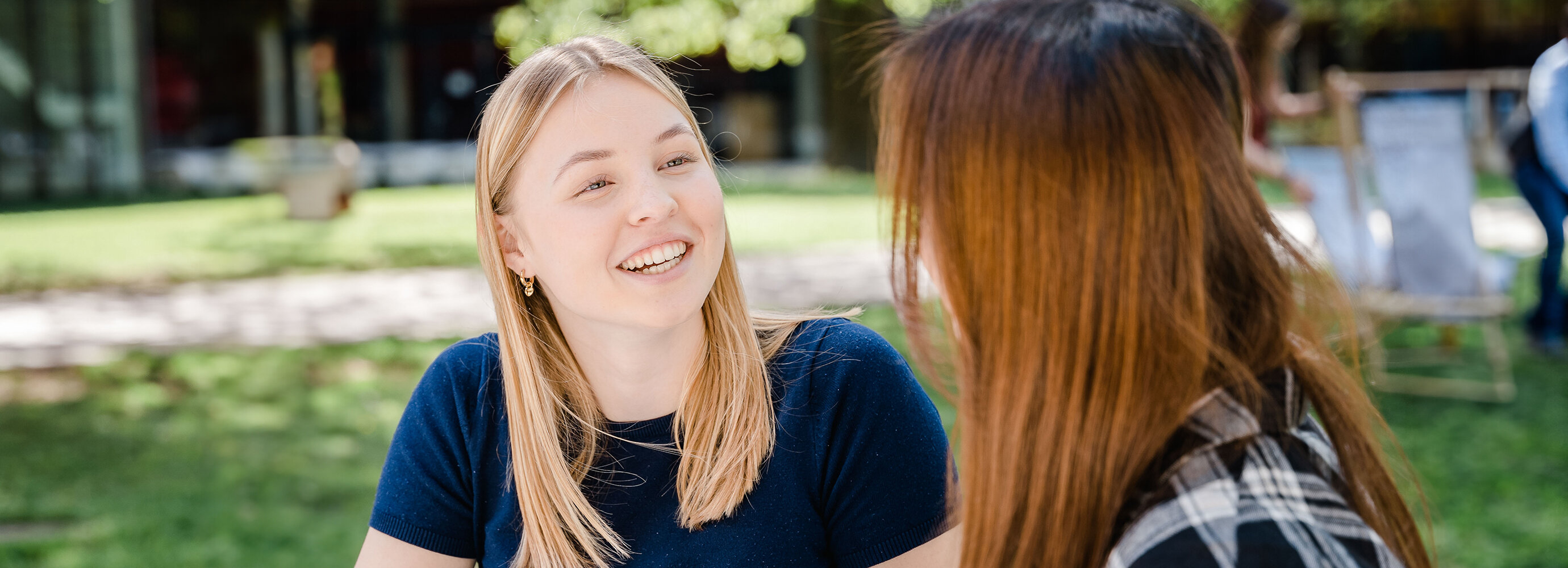 Zwei Frauen sitzen im Park der Hochschule und sprechen lächelnd miteinander. | © FH Kufstein Tirol