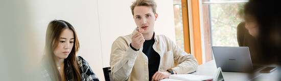 Drei Personen sitzen an einem Tisch, und ein Mann dieser Gruppe zeigt mit seinem Stift in die Richtung der Kamera, wo sich ein Whiteboard befindet. | © FH Kufstein Tirol