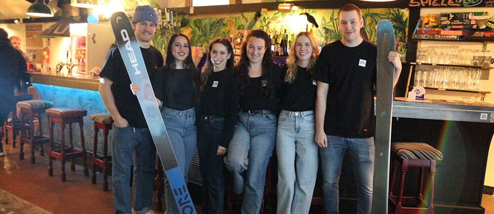 6 Personen stehen in Jeans und schwarzen T-Shirts vor einer Bar. Zwei haben jeweils einen Ski in der Hand. | © FH Kufstein Tirol