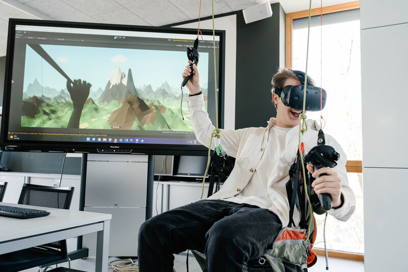 Web_Lab_VR_Simulation_1 Ein Mann sitzt in einer Schaukel und verwendet ein VR Headset mit 2 Controller.