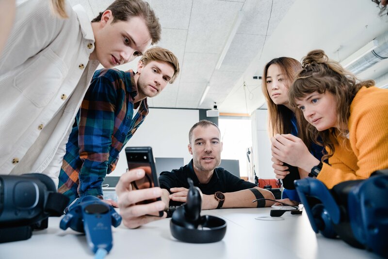 Web_Lab_Demonstration Ein Mann präsentiert etwas auf seinem Handy, während 4 andere Personen aufmerksam zuschauen.