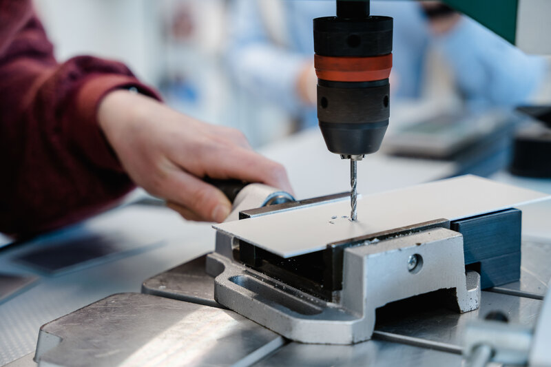 LEAN_Lab_Bohrmaschine_1 Eine Person bedient eine stationäre Bohrmaschine um Löcher in ein Metallwerkstück zu bohren.