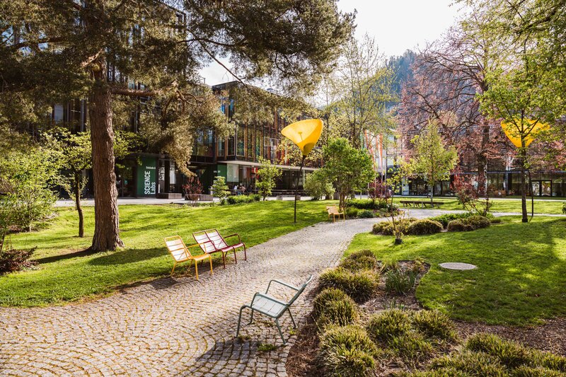 Idyllischer Außenbereich der FH Kufstein Tirol – Entspannen und Lernen im Grünen Grüner Campusbereich mit gepflastertem Weg, bunten Bänken und moderner Hochschularchitektur im Hintergrund