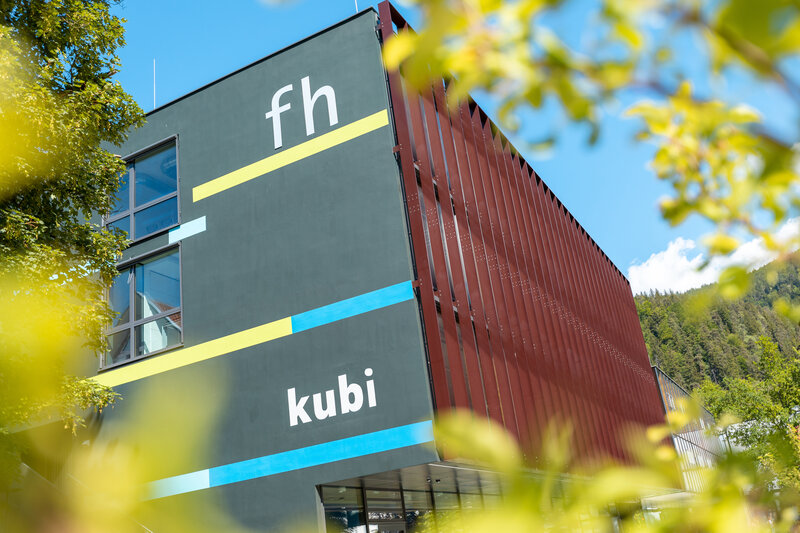 Das Gebäude der kubi in mitten des Kufsteiner Stadtparks Gebäude der FH Kufstein Tirol mit Schriftzug 'fh' und 'kubi' an der Fassade, umgeben von grünen Blättern und blauem Himmel.