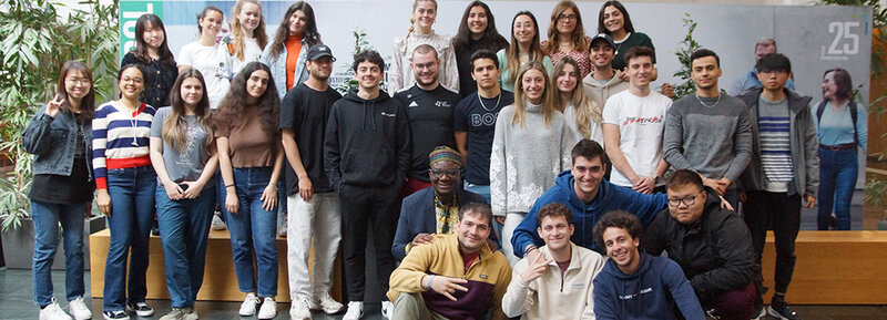 International_3 Eine Gruppe von Studierenden der FH Kufstein Tirol steht und sitzt lächelnd zusammen mit Gastlektor Bella Bello Bitugu in der Aula der Fachhochschule Kufstein Tirol.