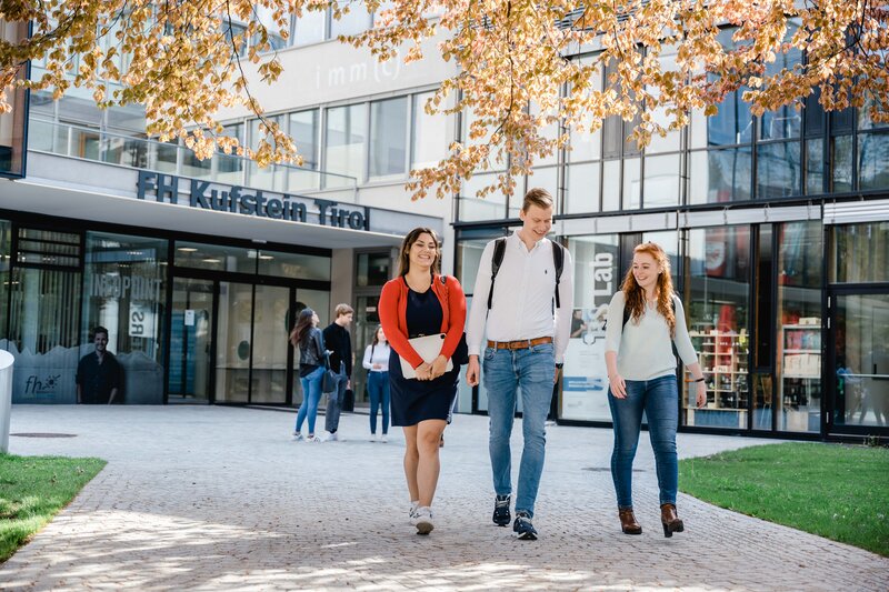 Park_Trio_gehend_1 Drei Personen gehen vom Eingang der FH Kufstein Tirol über den Steinpfad in Richtung Stadtpark.