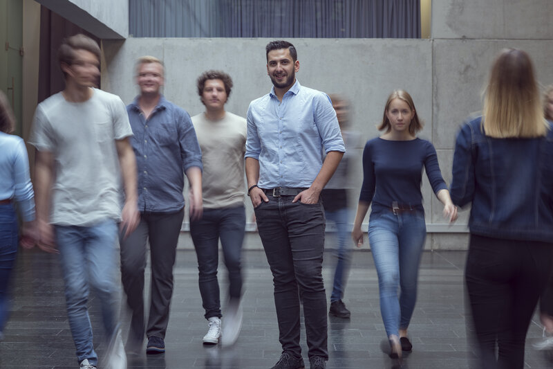 Aula_belebt_1 Viele Personen bewegen sich durch die Aula der FH Kufstein Tirol, und im Zentrum des Bildes steht ein Mann und blickt in die Kamera.