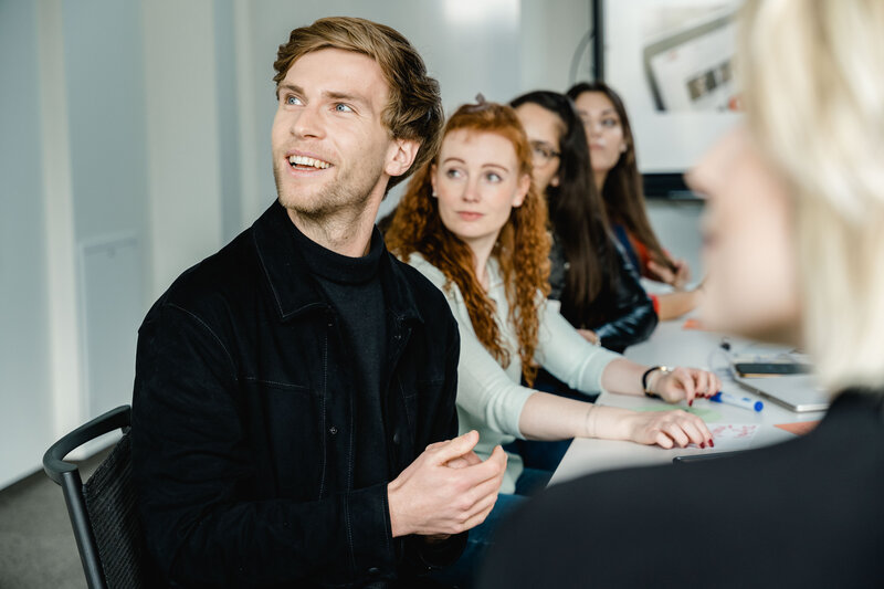 Meetingroom_Besprechung_1 Fünf Personen sitzen an einem Tisch, und blicken gespannt auf etwas außerhalb des Bildes.