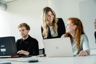 Arbeiten_am_Laptop Drei Personen sitzen an einem Tisch und arbeiten an ihren Laptops, während eine stehende Frau etwas mit einer sitzenden Frau bespricht.