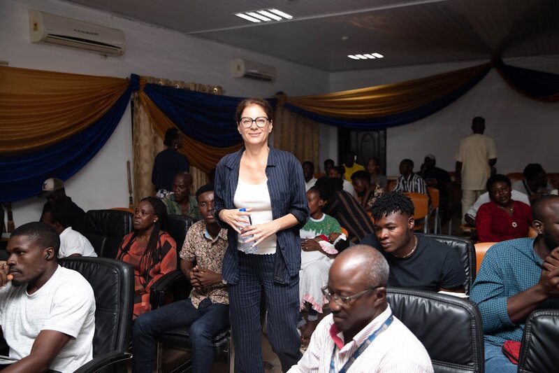 Ghana_Veranstaltung Eine Frau steht lächelnd in einem Raum voller Menschen, die auf Stühlen sitzen. Einige der sitzenden Personen unterhalten sich, während andere nach vorne schauen.