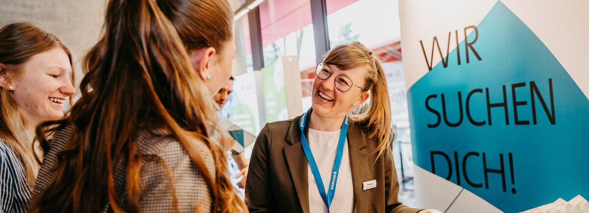 Meet & Match Drei Frauen im Gespräch an einem Messestand mit der Aufschrift "Wir suchen Dich!" im Hintergrund.