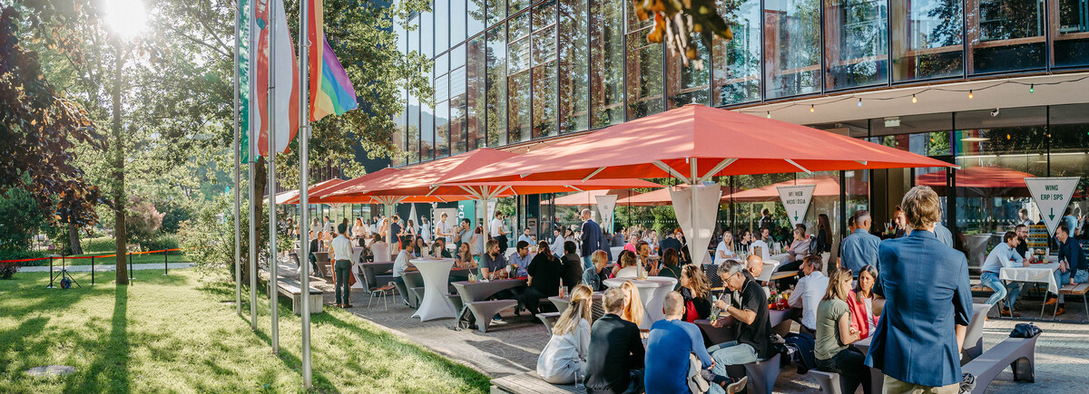 Home Coming 2024 Home Coming an der FH Kufstein Tirol. Festliche Veranstaltung. Menschen sitzen unter roten Sonnenschirmen in einem sonnigen Park neben einem modernen Glasgebäude. Die Atmosphäre ist entspannt und gesellig.