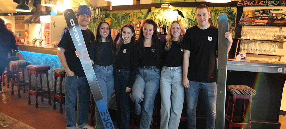 Event Team Peak & Powder Movie Night 2025 | © FH Kufstein Tirol 6 Personen stehen in Jeans und schwarzen T-Shirts vor einer Bar. Zwei haben jeweils einen Ski in der Hand. | © FH Kufstein Tirol