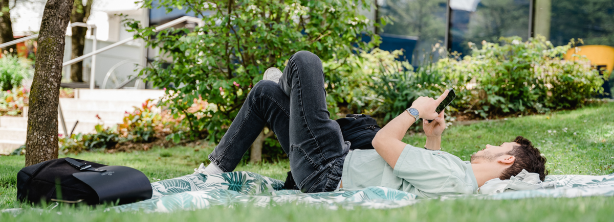Ein Mann liegt entspannt im Park auf einer Decke, und schaut in sein Smartphone. | © FH Kufstein Tirol Ein Mann liegt entspannt im Park auf einer Decke, und schaut in sein Smartphone. | © FH Kufstein Tirol